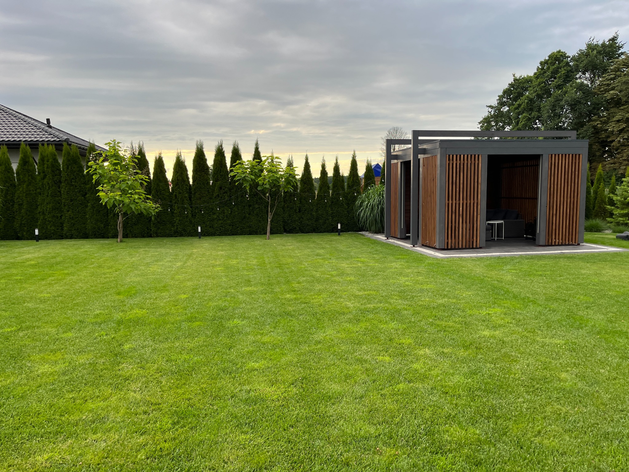 Nowoczesna, drewniana pergola z paletami na trawniku, otoczona tujami i młodymi drzewkami, pod pochmurnym niebem. Widoczny fragment dachu domu.