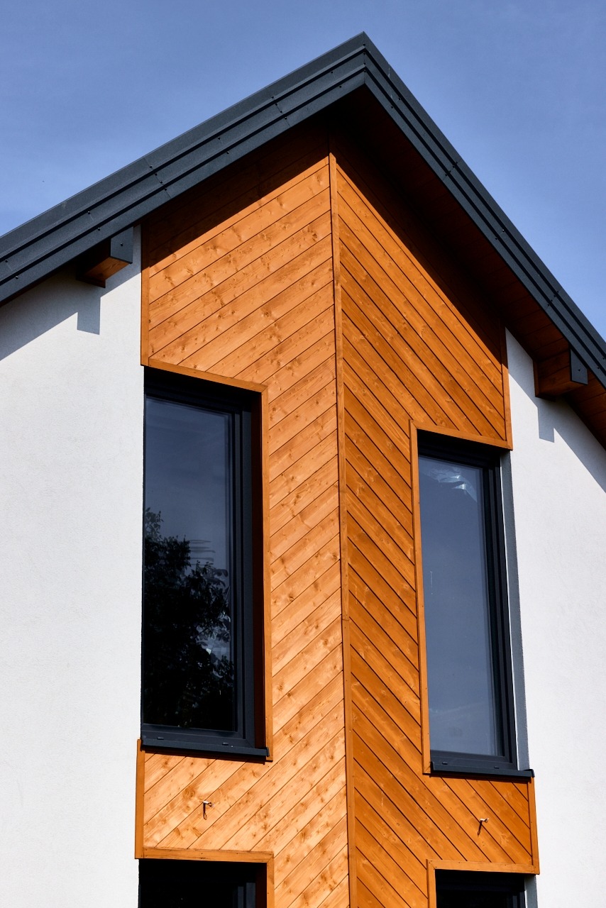 Fragment elewacji budynku z drewnianą okładziną ułożoną w jodełkę, widoczne dwa okna z czarnymi ramami i fragment dachu na tle błękitnego nieba.