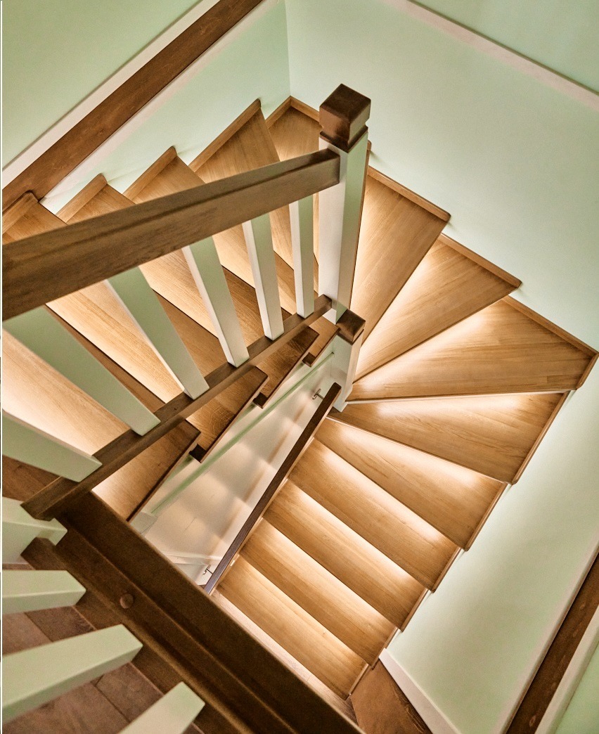 Drewniane schody zabiegowe z podświetleniem LED, widok z góry. Brązowe poręcze z białymi słupkami na tle jasnych ścian.