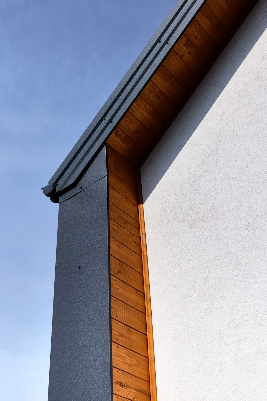 Fragment elewacji domu z drewnianym wykończeniem i metalową obróbką blacharską na tle błękitnego nieba w słoneczny dzień.