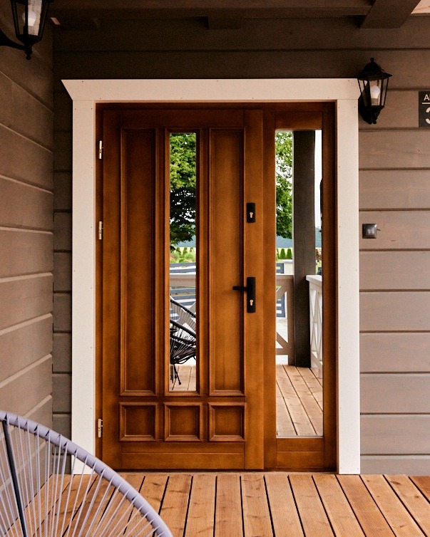 Drewniane drzwi wejściowe z przeszkleniami, osadzone w białej ramie, na tle szarej elewacji z drewnianą podłogą. Widoczny fragment krzesła.