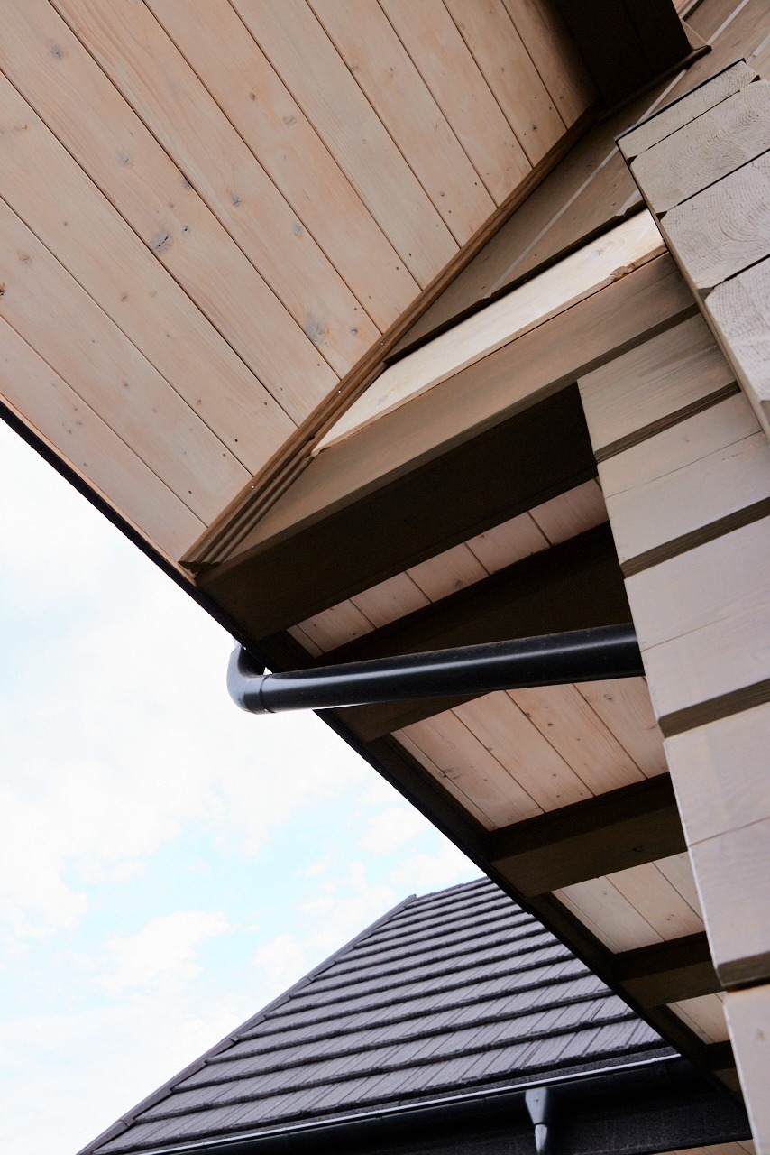 Fragment elewacji drewnianej z rynną w kolorze czarnym, widok z dołu na tle nieba. Widoczny fragment dachu z ciemną dachówką.
