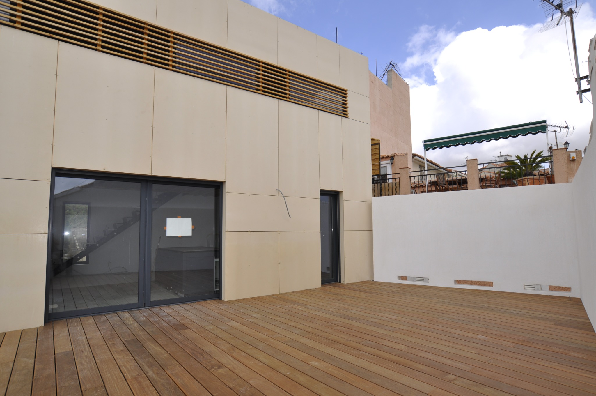 Nowoczesny balkon z drewnianą podłogą i jasną elewacją z poziomymi żaluzjami. Widoczne drzwi balkonowe i fragment sąsiedniego budynku.