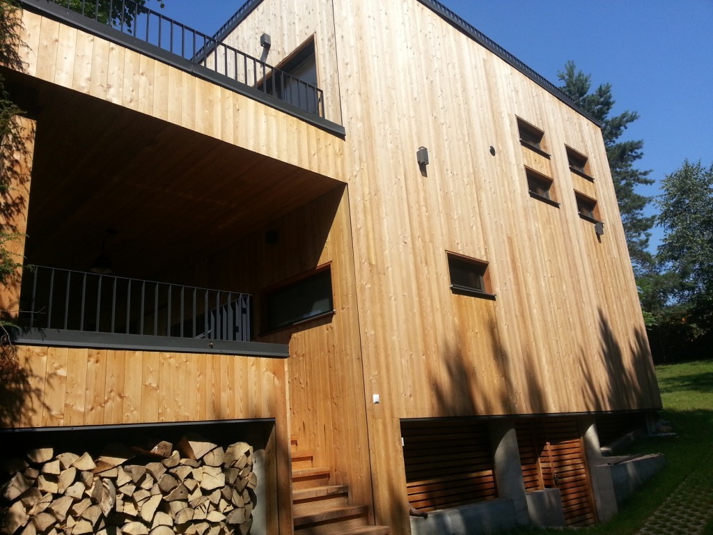 Nowoczesny dom z elewacją z jasnego drewna, widok z dołu. Elementy metalowe w kolorze czarnym. Składowane drewno pod balkonem. Minimalistyczny styl.