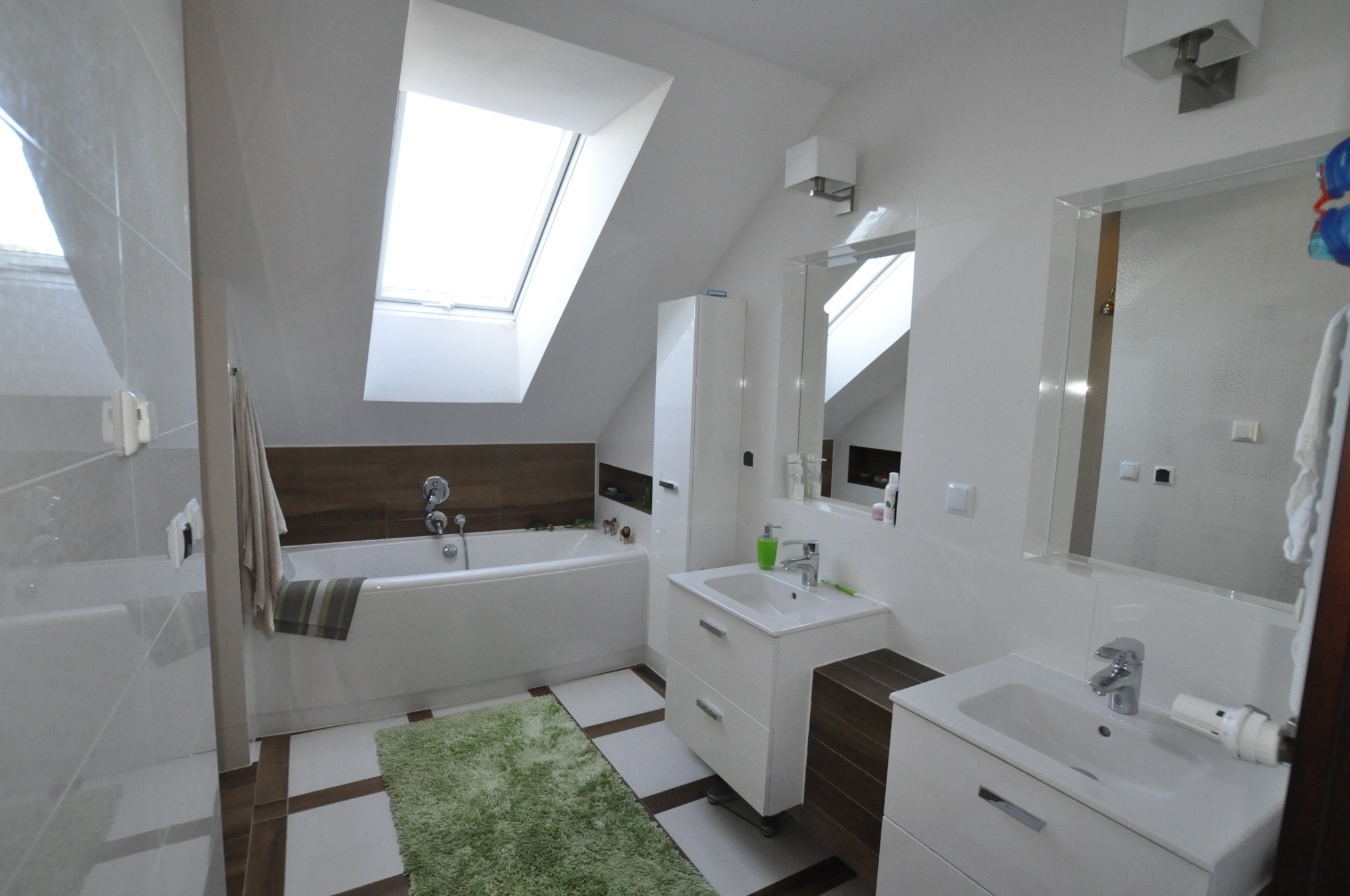 Jasna łazienka na poddaszu z oknem dachowym nad wanną. Białe meble łazienkowe, brązowe akcenty na podłodze i ścianie. Zielony dywanik dodaje koloru.