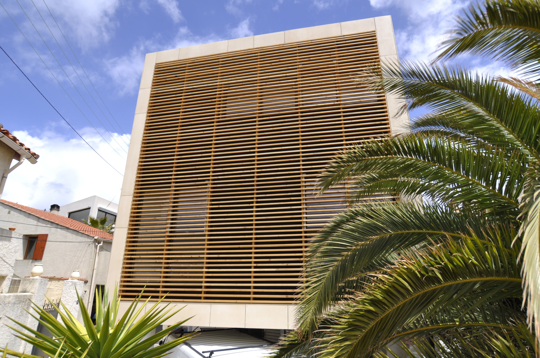 Nowoczesna elewacja budynku z drewnianymi żaluzjami, widziana z dołu, z fragmentami roślinności i błękitnym niebem w tle. Minimalistyczny design.
