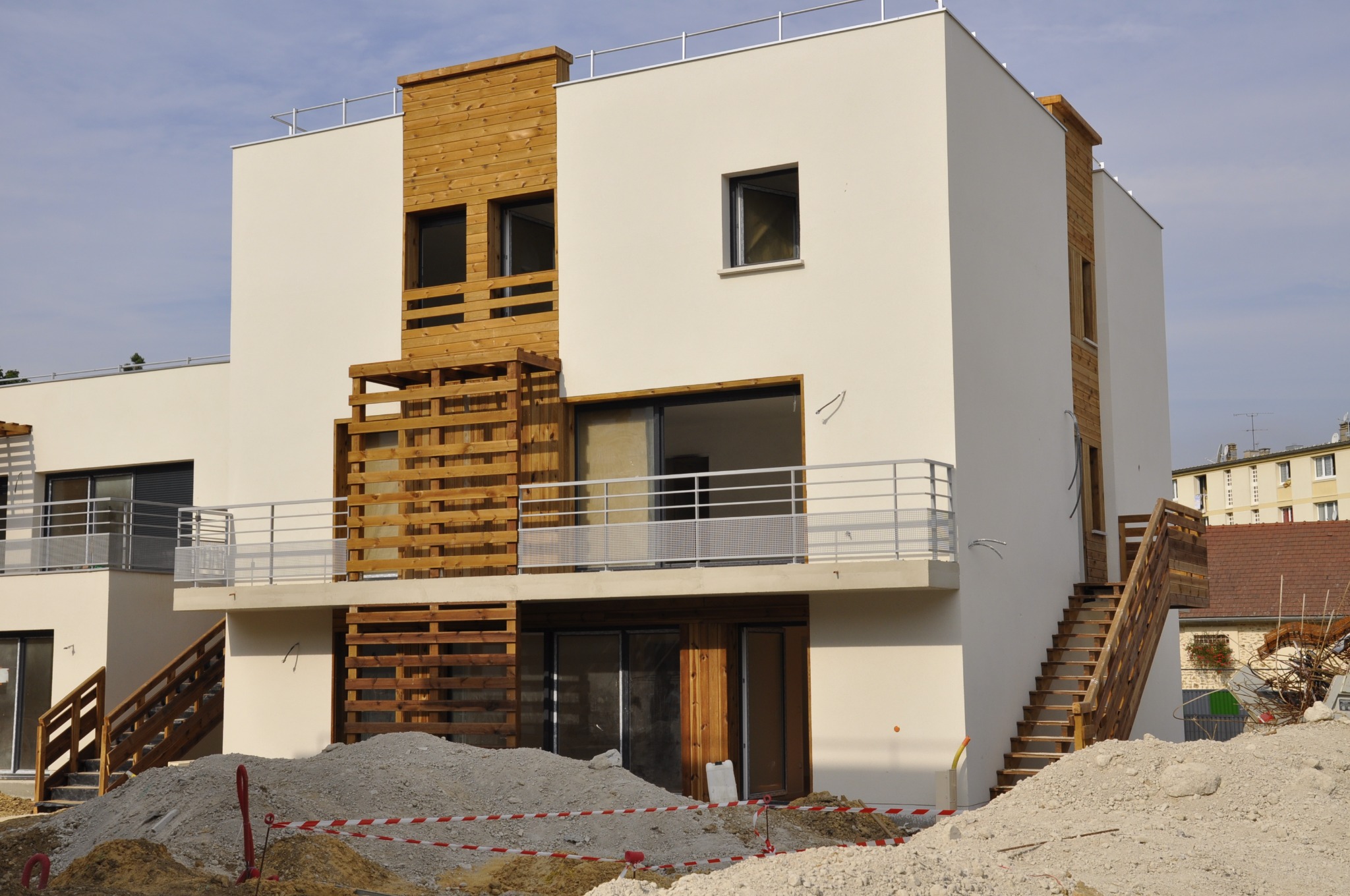Nowoczesny budynek z elewacją w odcieniach bieli i drewna, z balkonami i zewnętrznymi schodami. Widoczne materiały budowlane i taśmy ostrzegawcze.
