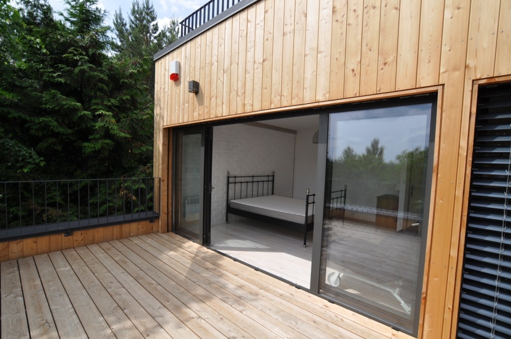 Drewniana elewacja domu z wyjściem na taras z desek. Widoczne przesuwne drzwi balkonowe z czarną ramą i fragment wnętrza z łóżkiem. Zewnętrzna barierka metalowa.