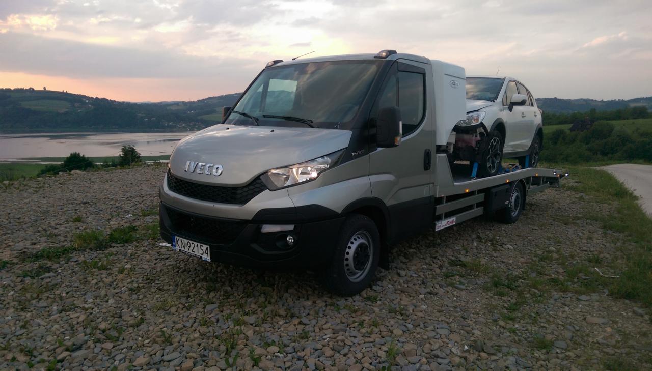 Srebrna laweta Iveco z uszkodzonym białym samochodem osobowym na platformie, zaparkowana na żwirowym podłożu z widokiem na jezioro i wzgórza w tle, o zmierzchu.