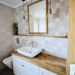 Jasna łazienka z drewnianym blatem, białą umywalką nablatową, lustrem w drewnianej ramie i wzorzystymi płytkami na ścianie. Wiszące lampy z żarówkami Edisona po bokach lustra.