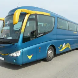 Niebieski autokar Irizar z żółtymi akcentami na nadwoziu i charakterystycznymi uchwytami nad przednią szybą, zaparkowany na betonowym placu.