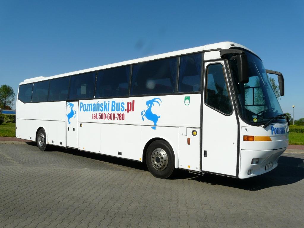 Biały autokar z niebieskim logo 'Poznański Bus.pl' z sylwetką koziołka na boku, zaparkowany na szarym bruku w słoneczny dzień.