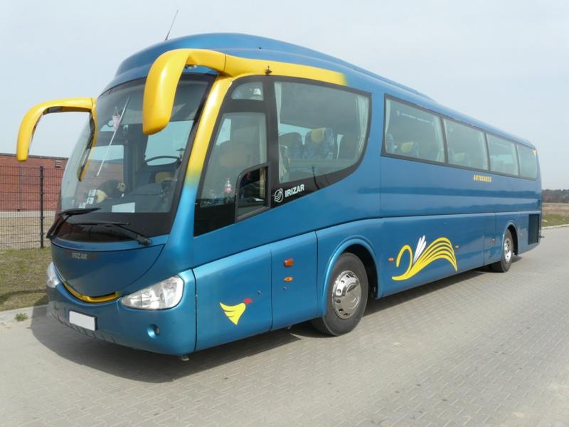 Niebieski autokar Irizar z żółtymi akcentami na nadwoziu i charakterystycznymi uchwytami nad przednią szybą, zaparkowany na betonowym placu.