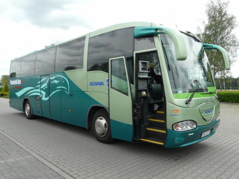 Zielono-turkusowy autokar turystyczny Scania z otwartymi drzwiami i logo firmy przewozowej na boku, zaparkowany na brukowanym placu.