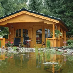 Drewniany domek letniskowy z zadaszonym tarasem, odbijający się w stawie, otoczony zielenią i kwiatami, z dwoma krzesłami na tarasie.