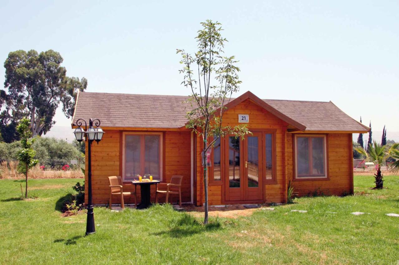 Mały, parterowy domek z bali z numerem 21 na ścianie, ustawiony na zadbanym trawniku, z widoczną lampą ogrodową i stolikiem z dwoma krzesłami przed oknem.