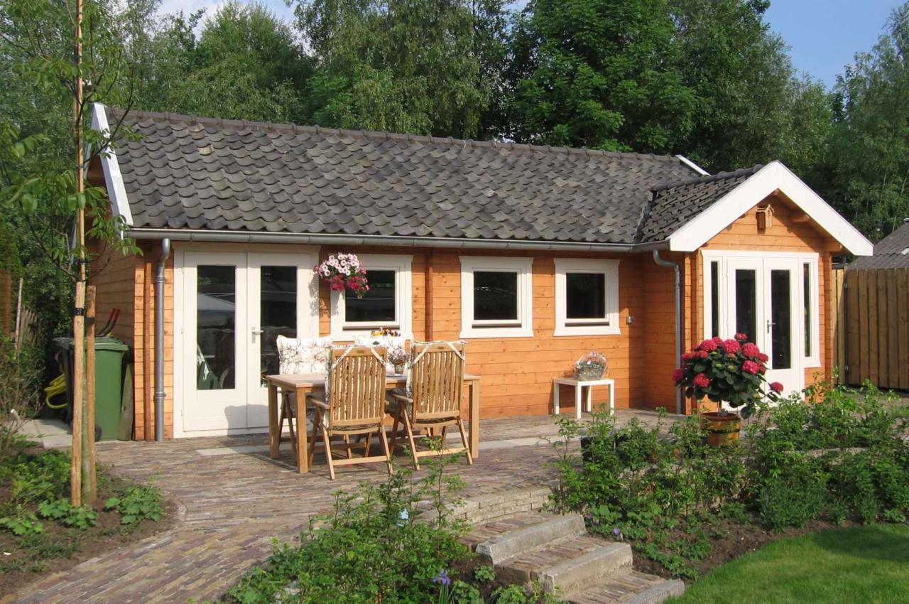 Drewniany domek z szarym dachem, białymi oknami i drzwiami, z drewnianym stołem i krzesłami na brukowanym patio, otoczony zielenią.