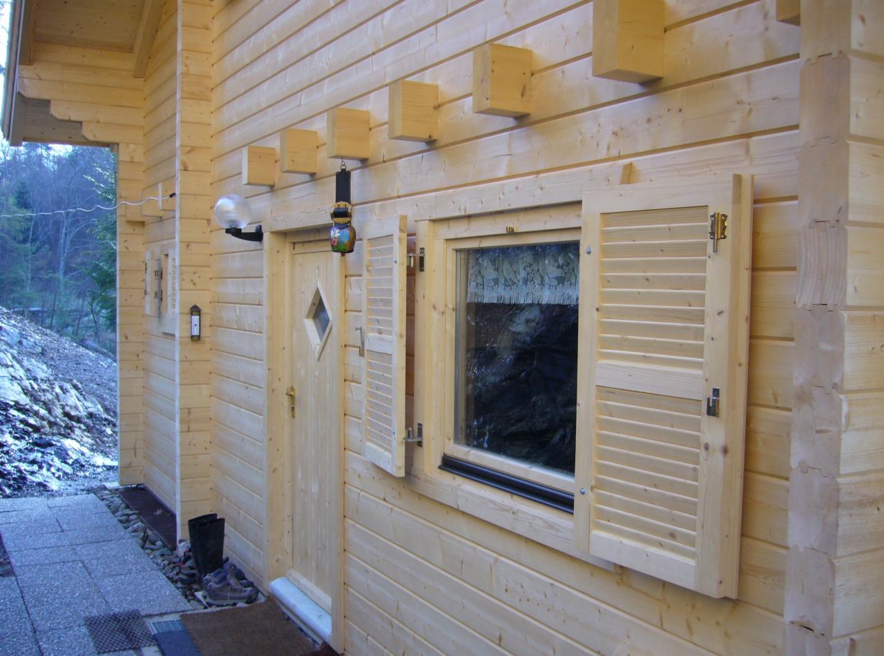 Zewnętrzny widok na drewniany dom z bali z oknem z okiennicami, drzwi z romboidalnym oknem i dekoracyjnym dzwonkiem nad drzwiami, oświetlenie zewnętrzne i kamienną ścieżką prowadzącą do wejścia.