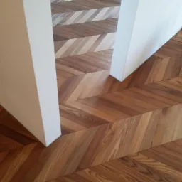 Parkiet ułożony we wzór jodełki francuskiej, z widocznymi różnicami w odcieniach drewna, przylegający do białej ściany.