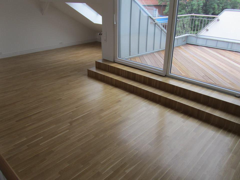 Jasna dębowa podłoga w pokoju z wyjściem na taras, trzy stopnie prowadzą do przeszklonych drzwi balkonowych, widoczny fragment tarasu z drewnianą podłogą i metalową balustradą.