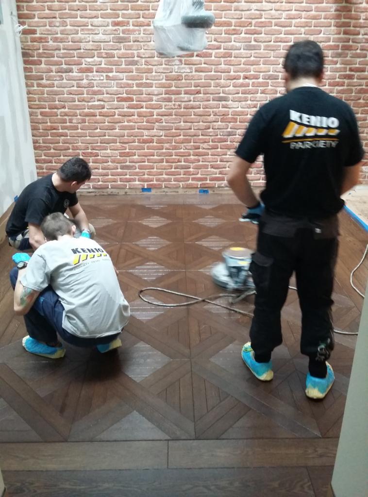 Parkieciarze w trakcie cyklinowania parkietu o wzorze szachownicy w pomieszczeniu z ceglaną ścianą, widoczna maszyna do cyklinowania i logo firmy na koszulkach.