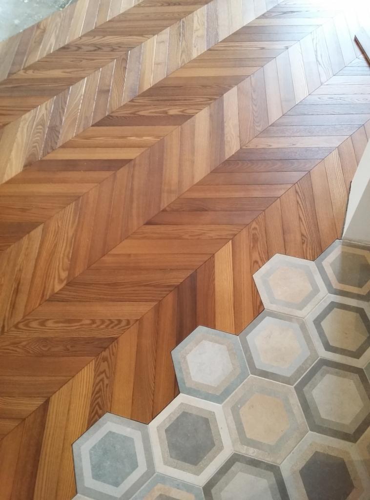 Jodełka z parkietu dębowego przechodzi w heksagonalne płytki ceramiczne w odcieniach szarości i beżu, widok z góry.