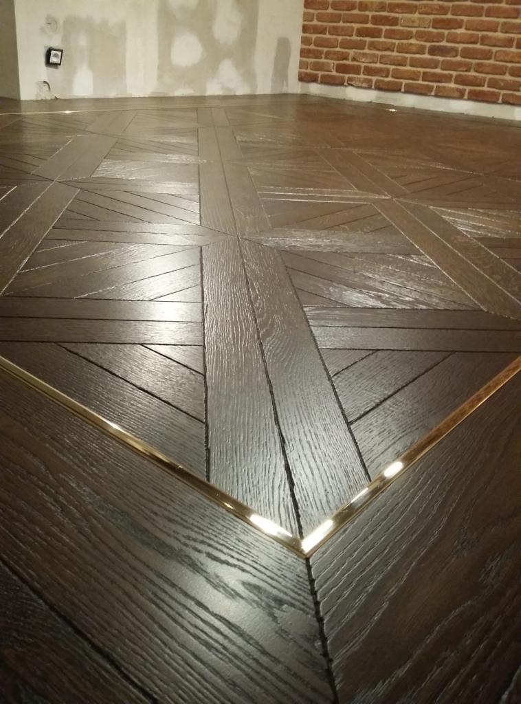 Ciemna drewniana podłoga ułożona we wzór kwadratów z metalowymi listwami wykończeniowymi, ściana z cegły w tle.
