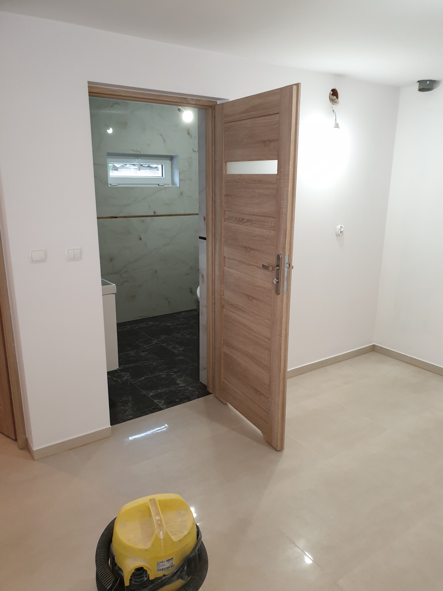 Wnętrze po remoncie: widok na łazienkę z marmurowymi płytkami, otwarte drewniane drzwi, żółty odkurzacz na jasnych płytkach podłogowych, niedokończone gniazdko elektryczne na ścianie.
