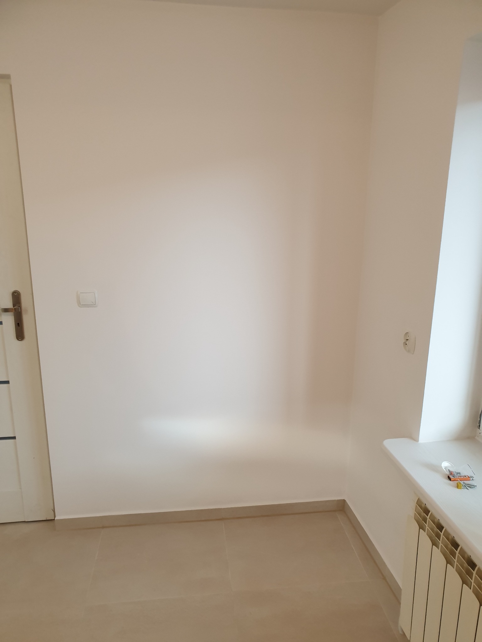 Pomalowane na jasny beż narożne wnętrze pokoju z grzejnikiem pod oknem, widoczne narzędzia na parapecie i fragment drzwi z klamką.