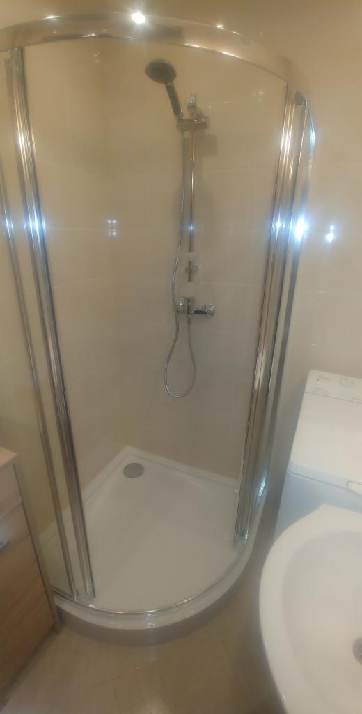 Kabiną prysznicowa z chromowanymi profilami i deszczownicą, widoczna umywalka i pralka w tle, beżowe płytki na ścianach.