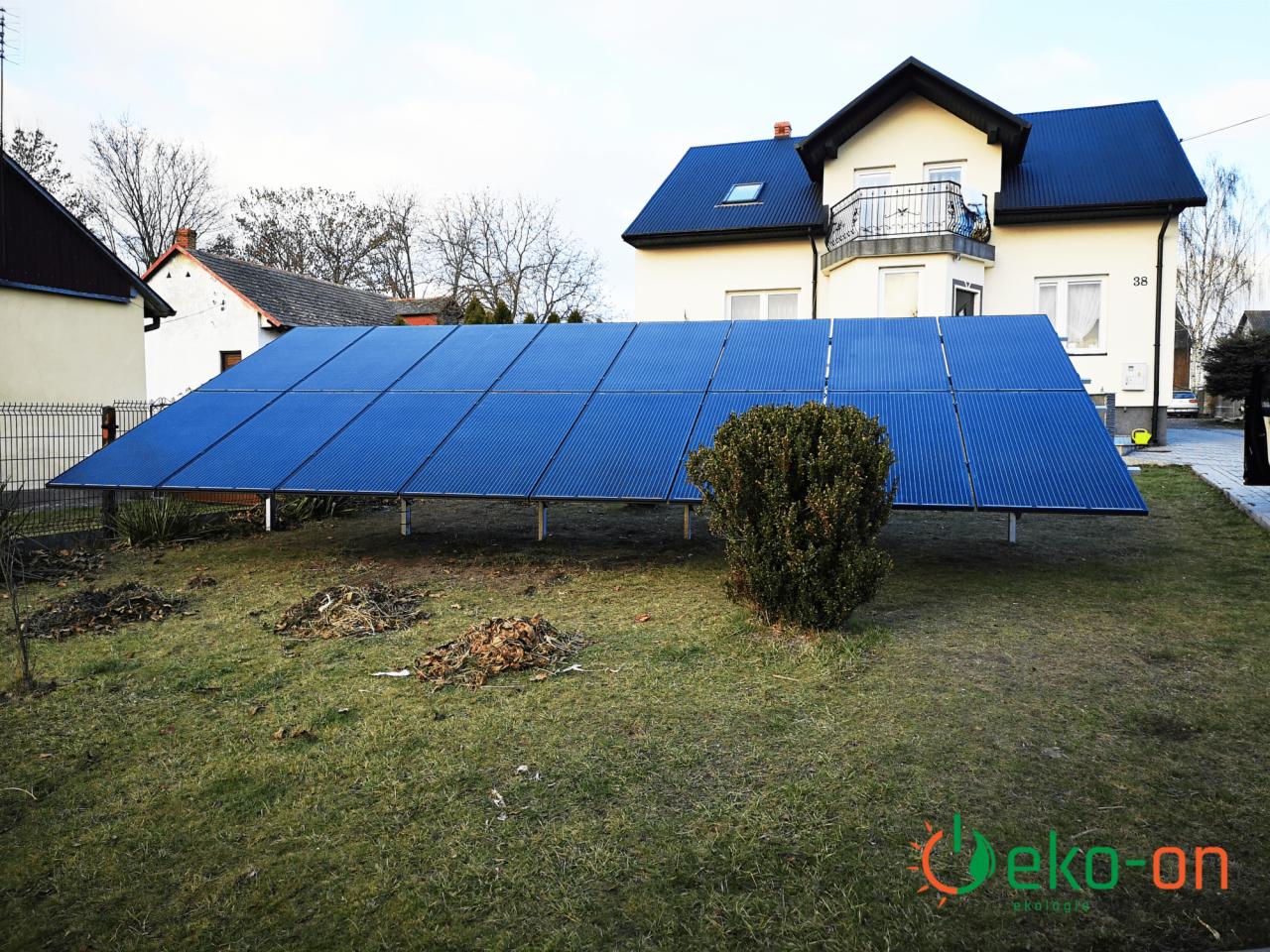 Instalacja paneli słonecznych na niskim stelażu na trawniku przed domem z balkonem, widoczny krzew i sterta liści.