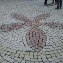 Wzór kwiatowy ułożony z kostki granitowej w odcieniach szarości i brązu, widoczne fragmenty nóg przechodniów w tle.