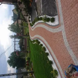 Falujący chodnik z czerwonej i szarej kostki brukowej, obrzeża z białego grysu, ozdobne krzewy, trawnik, huśtawka ogrodowa w tle.