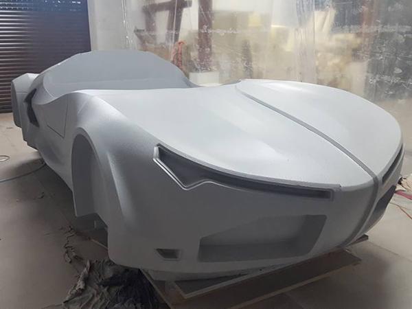 Szary prototyp sportowego samochodu Hydrocar Premier na drewnianej palecie, widoczne zarysowane reflektory i linia podziału maski, w tle folia ochronna.