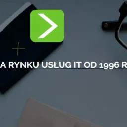 Biurko z elementami związanymi z IT: notatnik, logo firmy IT, okulary, ołówek i roślina doniczkowa. Widoczny napis 'Działamy na rynku usług IT od 1996 roku'.