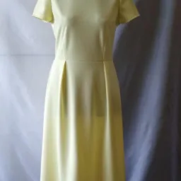 Żółta, dopasowana sukienka z krótkim rękawem i plisami w talii, prezentowana na manekinie na tle białej i szarej tkaniny.