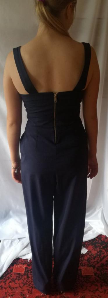 Tył granatowego damskiego kombinezonu z szerokimi nogawkami i złotym zamkiem błyskawicznym na plecach, na tle białej tkaniny i czerwonego dywanu.