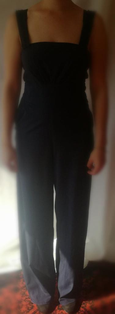 Osoba w czarnym kombinezonie z szerokimi nogawkami i grubymi ramiączkami na tle ściany i wzorzystego dywanu.