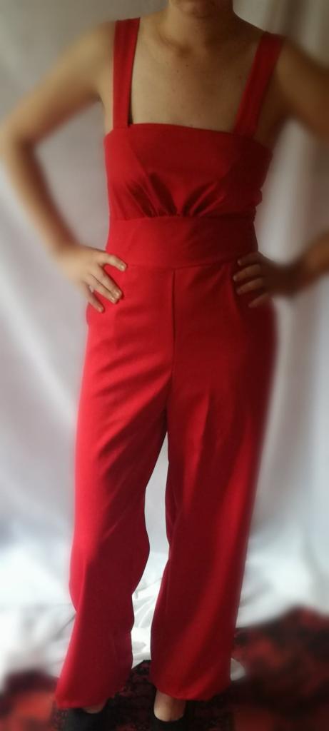 Osoba prezentuje czerwony kombinezon z szerokimi nogawkami i kwadratowym dekoltem na tle białej tkaniny.
