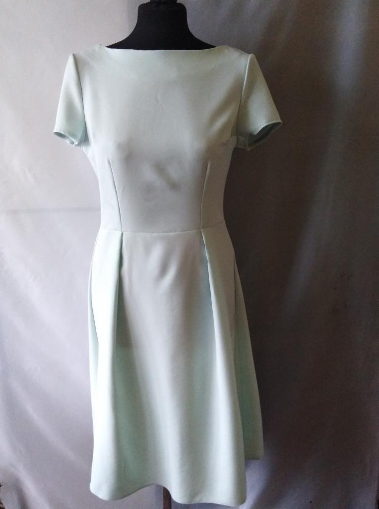 Jasnozielona, minimalistyczna sukienka z krótkim rękawem i plisowanym dołem, prezentowana na manekinie na tle szarej i białej tkaniny.