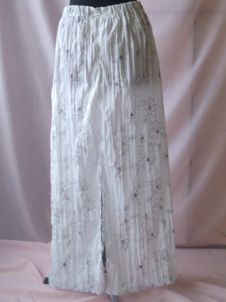 Długa, biała, plisowana spódnica z rozcięciem i delikatnym kwiatowym wzorem prezentowana na manekinie w Rędzinach, na tle różowej tkaniny.