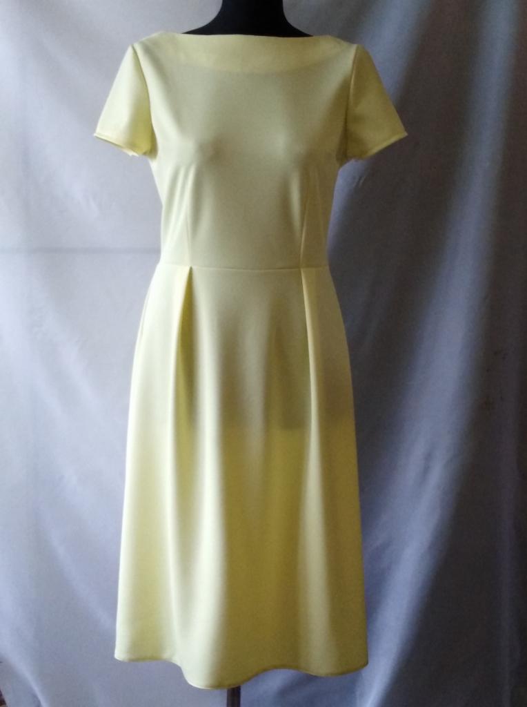 Żółta, dopasowana sukienka z krótkim rękawem i plisami w talii, prezentowana na manekinie na tle białej i szarej tkaniny.