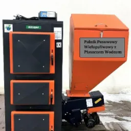 Wielopaliwowy piec CO z podajnikiem ślimakowym i zasobnikiem na paliwo w kolorze pomarańczowym, widoczne trzy drzwiczki paleniska i sterownik elektroniczny.