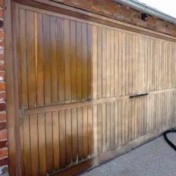 Drewniana brama garażowa częściowo oczyszczona z zabrudzeń, ukazująca różnicę między stanem przed i po piaskowaniu, z widocznym wężem piaskarki leżącym na ziemi.