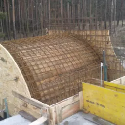 Szkielet łukowego elementu konstrukcyjnego zbrojony stalową siatką, oszalowany płytami drewnianymi, widoczne elementy szalunków Doka 3S basic, w tle las i teren budowy.