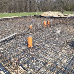 Siatka zbrojeniowa fundamentu z wystającymi rurami kanalizacyjnymi w kolorze pomarańczowym i szarym, zabezpieczona czarną folią, widok z góry na tle terenu budowy.