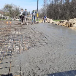 Robotnicy podczas wylewania betonu na zbrojoną siatką stalową, widoczna pompa do betonu i przygotowany teren budowy.