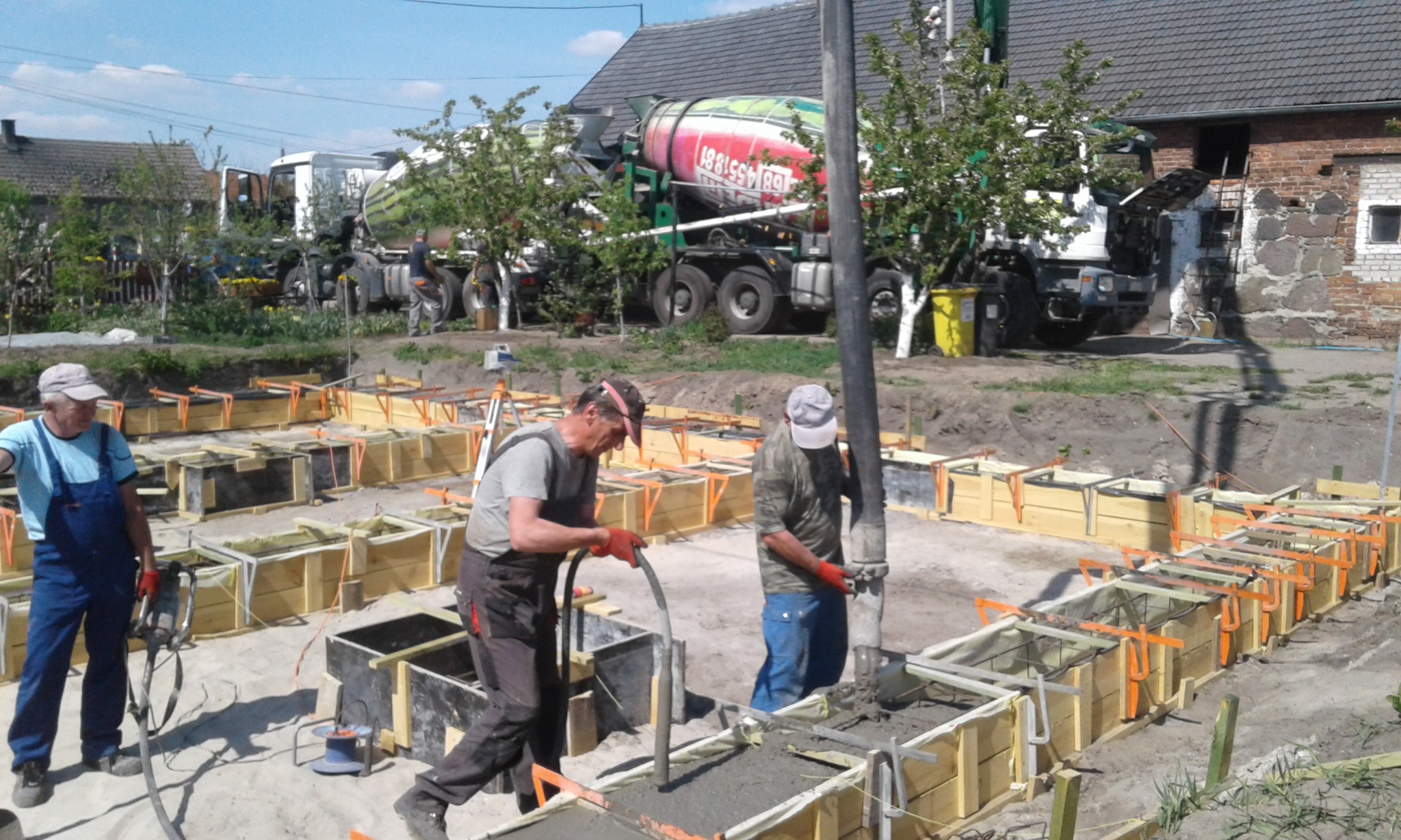 Wylewanie betonu do szalunków fundamentowych z użyciem pompy, z widocznymi zbrojeniami i pracownikami w trakcie pracy, na tle betonomieszarek i zabudowań.