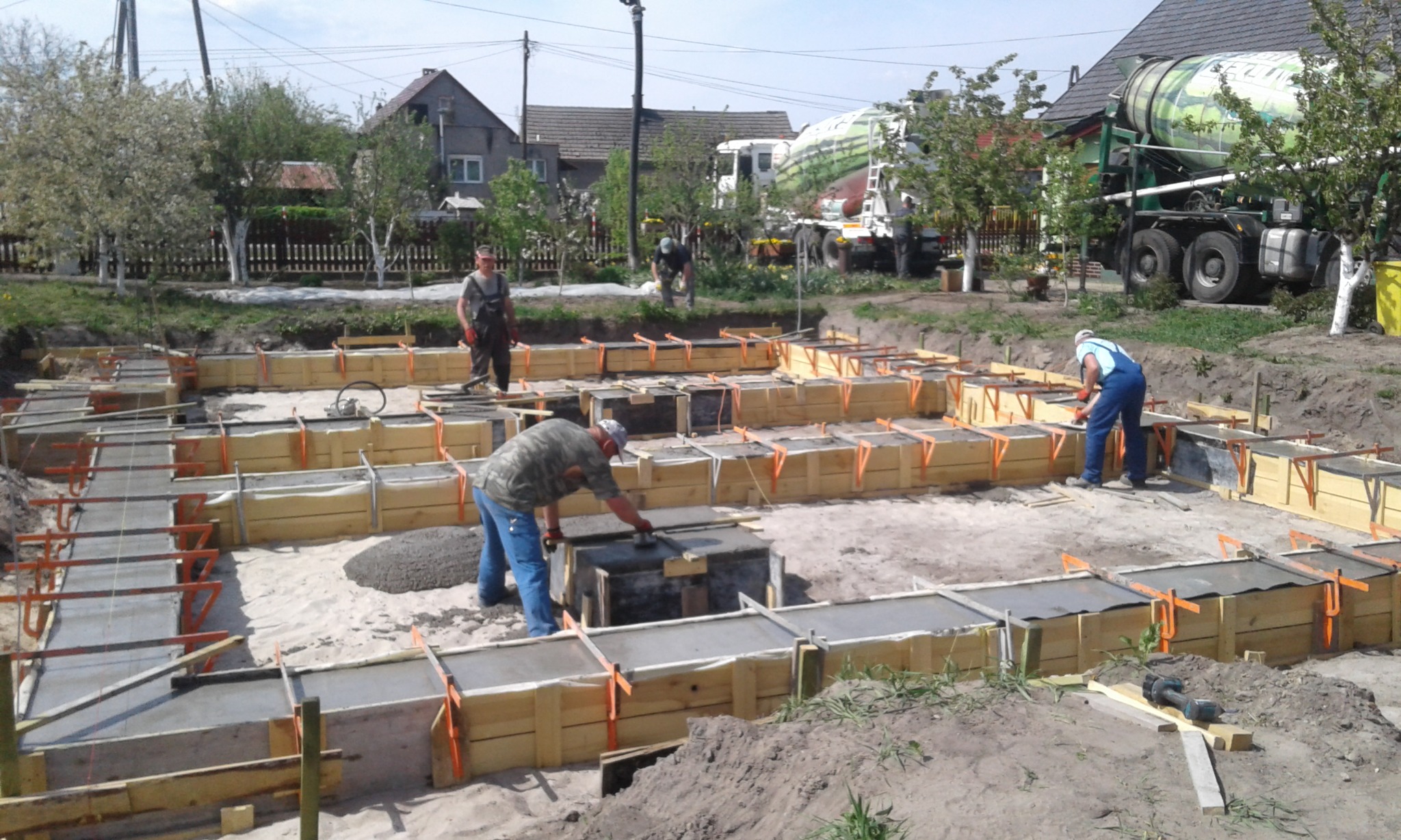 Wykonywanie fundamentów pod budynek: zalewanie betonem szalunków z drewnianych desek, widoczny betonowóz na działce.
