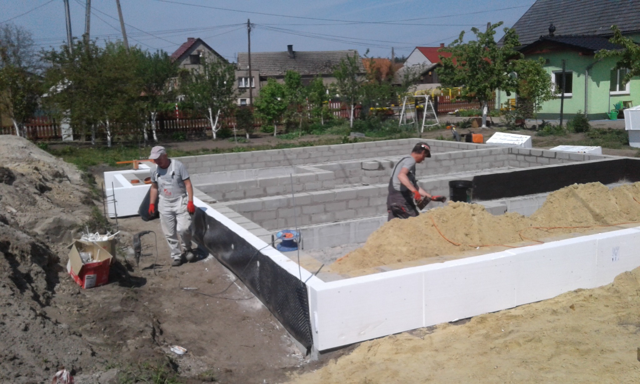 Dwóch robotników w trakcie budowy fundamentów domu z bloczków betonowych i białego styropianu, widoczne narzędzia i piasek, w tle zabudowa jednorodzinna.